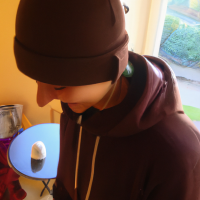 Pixar’s 3d style young man looking down with a white plush cap and brim front faced. Brown sweatshirt with hood and drawstring. Medium-sized beard all over the face. Shy laughter in the corner of his mouth. Gold hoop earring in the ear and black hoop piercing in the nose. Brown eyes and short hair. Skin is white and face is slightly rounded. Diagonal view facing left.