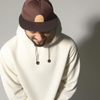 Pixar’s 3d style young man looking down with a white plush cap and brim front faced. Brown sweatshirt with hood and drawstring. Medium-sized beard all over the face. Shy laughter in the corner of his mouth. Gold hoop earring in the ear and black hoop piercing in the nose. Brown eyes and short hair. Skin is white and face is slightly rounded. Diagonal view facing left.