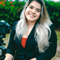 Fotógrafa, blazer preto, cabelos loiros um pouco abaixo dos ombros, sorriso fácil, com uma pinta no lado esquerdo do rosto, abaixo da boca